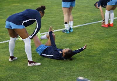 A group of athletes in sportswear are on a green soccer field. Two individuals are interacting closely; one helps the other who is lying on the grass. Nearby, other athletes are stretching.
