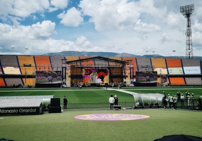 A sports stadium with a large stage set up on the field, hosting an outdoor event. Spectators wearing colorful hats sit in the foreground. The seating areas display vibrant colors, and a clear blue sky with scattered clouds is visible. Large screens display graphics related to the event.