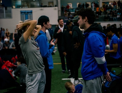 People engage in conversation and activities in an indoor sports facility. A woman in gray athletic wear stretches her arms above her head while talking to a man in a blue jacket. Other people are seen interacting in the background, some are seated on the ground, and the atmosphere is lively with many individuals gathered.