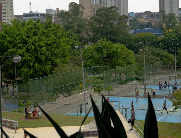 An outdoor recreational area in an urban setting featuring several sports courts and surrounded by lush green trees. People are engaged in various activities, including playing sports on the courts. In the background, there are tall buildings indicative of a cityscape.